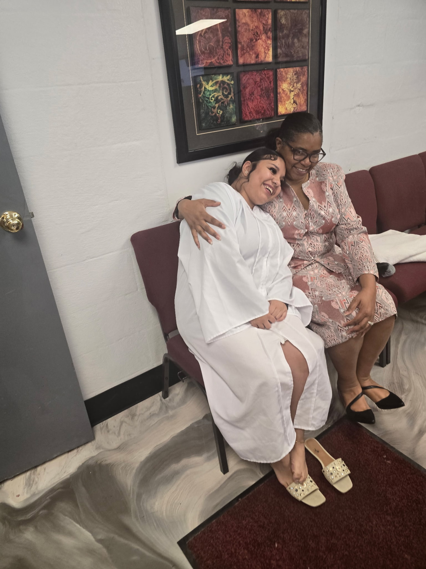 Young woman in a white baptismal robe embracing an older woman on a church pew